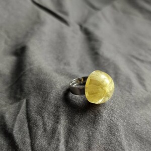 May include: A silver ring featuring a large, rounded, yellow-green gemstone. The gemstone has a textured, translucent appearance. The ring is set against a grey fabric background, highlighting the jewellery's design and colour.