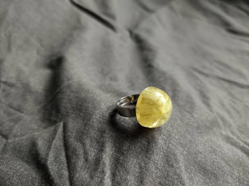 May include: A silver ring featuring a large, rounded, yellow-green gemstone. The gemstone has a textured, translucent appearance. The ring is set against a grey fabric background, highlighting the jewellery's design and colour.
