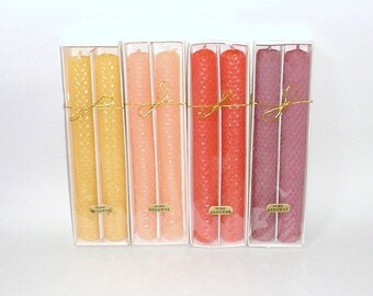 May include: Four sets of beeswax taper candles in clear boxes. Each set has two candles in shades of yellow, peach, red, and purple. The candles are tied with gold string and have small labels that read "Pure Beeswax".