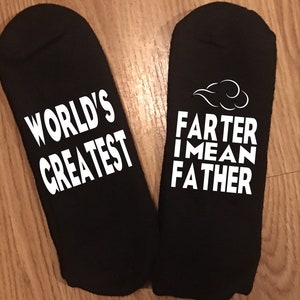 May include: Black socks with white text. One sock reads "WORLD'S GREATEST", the other reads "FARTER I MEAN FATHER" with a cloud graphic. The socks are likely a novelty gift.