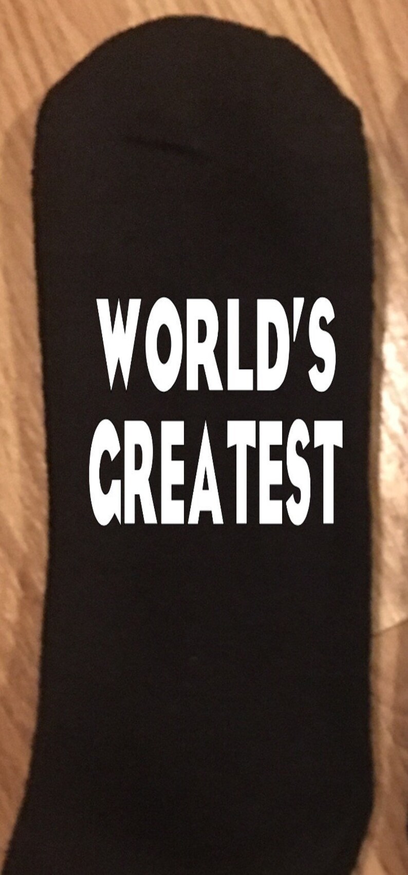 May include: Black sock with the words "WORLD'S GREATEST" in bold white letters. The sock is made of a soft material and is likely for everyday wear. The text is centered on the sock.