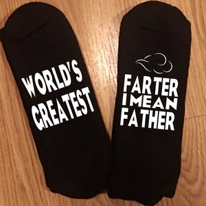 May include: A pair of black socks with white text. One sock reads "WORLD'S GREATEST", and the other reads "FARTER I MEAN FATHER" with a cloud graphic. The socks are displayed on a wooden surface.