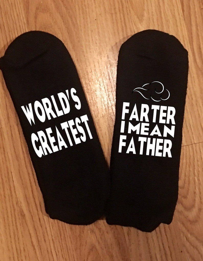 May include: A pair of black socks with white text. One sock reads "WORLD'S GREATEST", and the other reads "FARTER I MEAN FATHER" with a cloud graphic. The socks are displayed on a wooden surface.