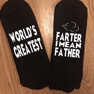 May include: A pair of black socks with white text. One sock reads "WORLD'S GREATEST" and the other reads "FARTER I MEAN FATHER" with a cloud graphic. The socks are displayed on a wooden surface.