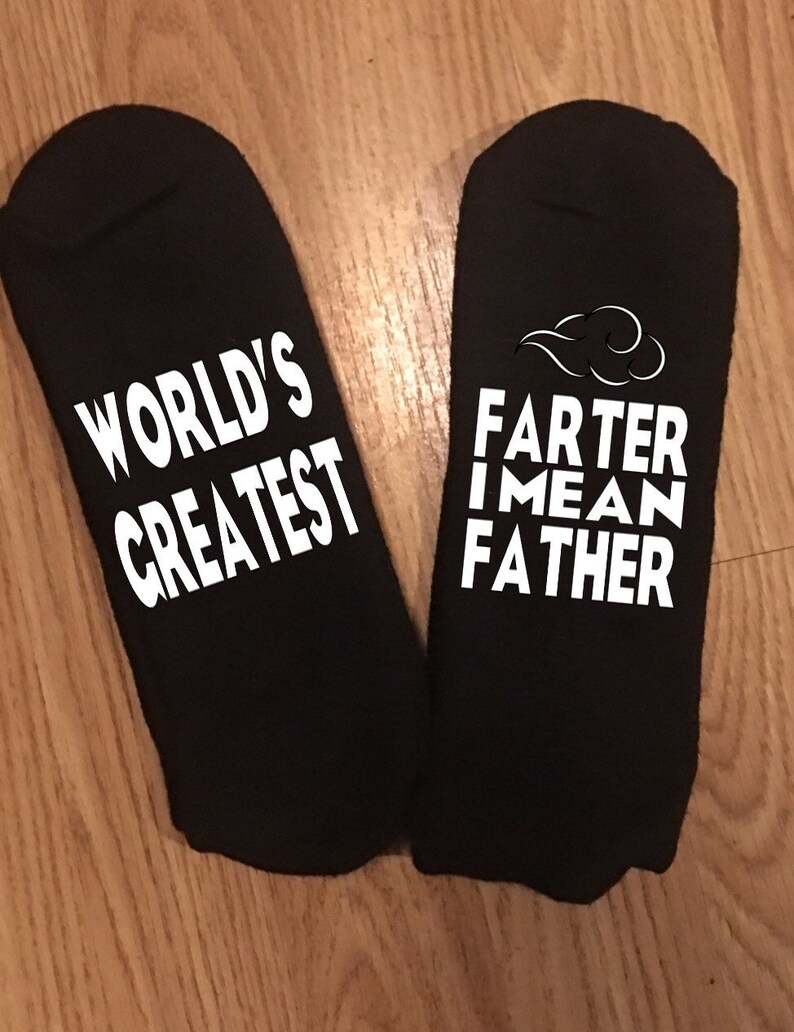 May include: A pair of black socks with white text. One sock reads "WORLD'S GREATEST" and the other reads "FARTER I MEAN FATHER" with a cloud graphic. The socks are displayed on a wooden surface.
