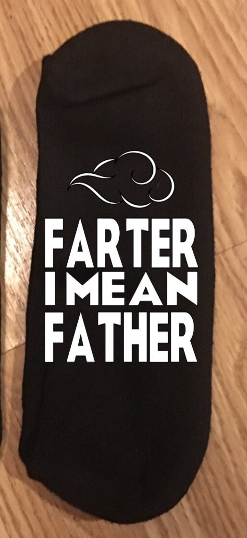 May include: Black sock with the text "FARTER I MEAN FATHER" in white block letters. Above the text is a white cloud-like design. The sock is laying on a wooden surface.