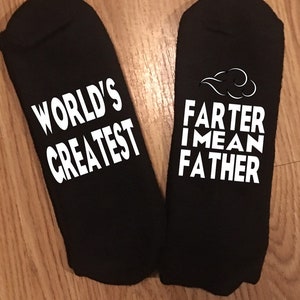 May include: A pair of black ankle socks with white text. One sock reads "WORLD'S GREATEST", the other reads "FARTER I MEAN FATHER" with a cloud graphic. The socks are displayed on a wooden surface.