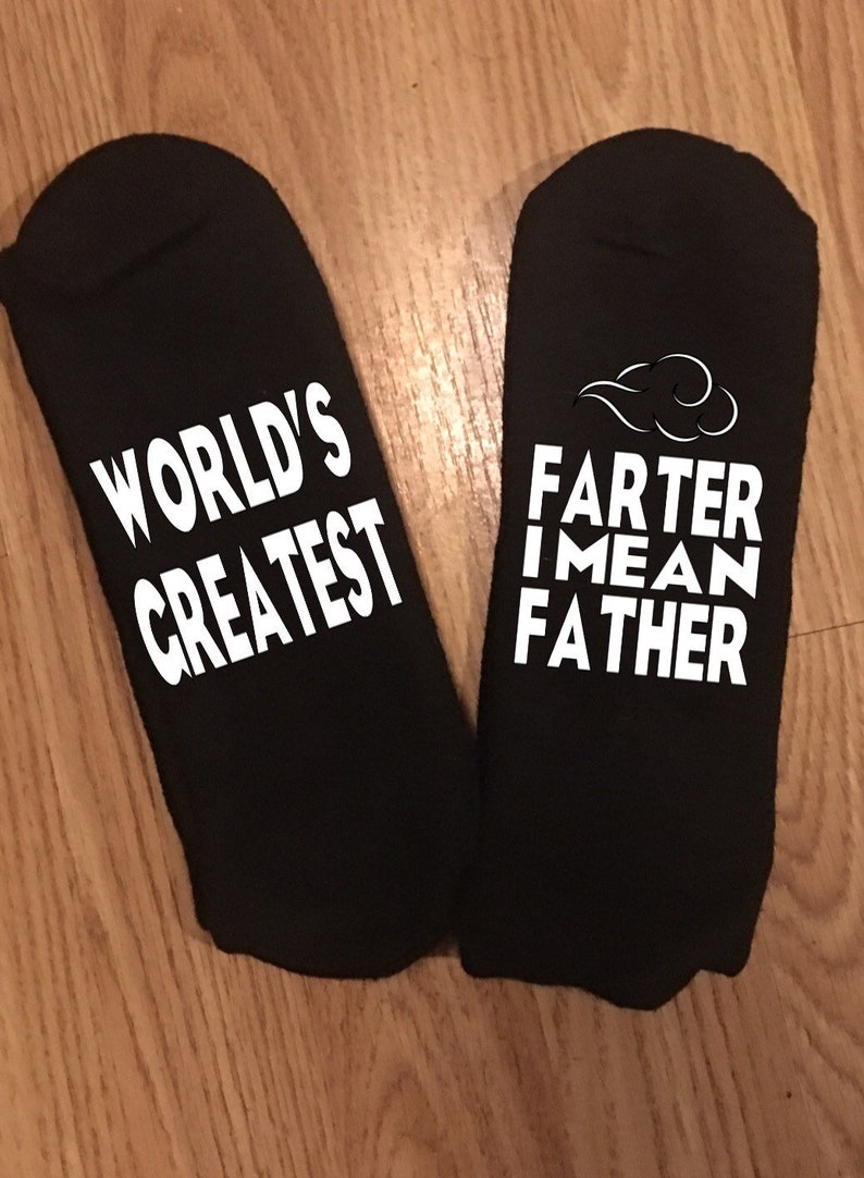 May include: A pair of black ankle socks with white text. One sock reads "WORLD'S GREATEST", the other reads "FARTER I MEAN FATHER" with a cloud graphic. The socks are displayed on a wooden surface.