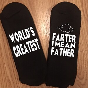May include: Two black socks with white text. One sock reads "WORLD'S GREATEST". The other sock reads "FARTER I MEAN FATHER" with a cloud graphic above the text. The socks are displayed on a wooden surface.