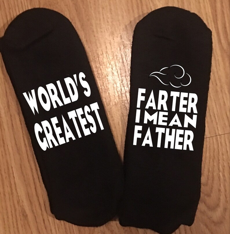 May include: Two black socks with white text. One sock reads "WORLD'S GREATEST". The other sock reads "FARTER I MEAN FATHER" with a cloud graphic above the text. The socks are displayed on a wooden surface.
