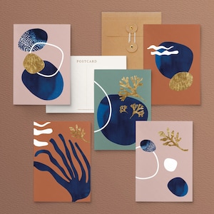 May include: A selection of abstract art postcards with diverse designs. The postcards showcase navy blue shapes, gold leaf accents, and white line details on a palette of muted colours, including pink, teal, and rust. A kraft paper envelope and a blank postcard are also included.