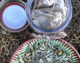Puede incluir: Un tarro de vidrio lleno de bolsitas de té de papel marrón se encuentra sobre un lecho de musgo verde. Un cuenco de cerámica marrón lleno de hierbas y flores secas está al lado del tarro.