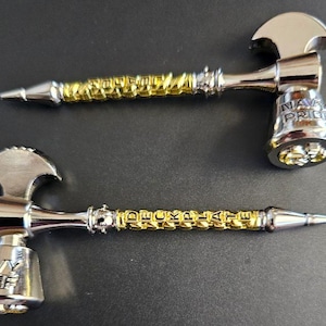 May include: Two silver and gold metal novelty items shaped like small axes. One reads "NAVY PRIDE" and the other reads "NAVY CHIEF". The handles are gold-colored and have text. The axes are on a dark surface.
