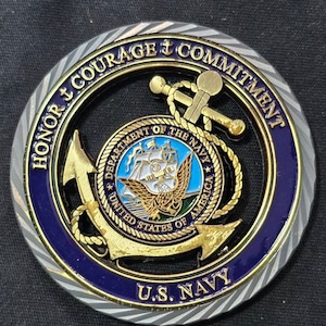 May include: A round U.S. Navy coin with a gold anchor, rope, and emblem. The coin features the words "Honor, Courage, Commitment" and "Department of the Navy, United States of America." The coin has a blue and gold color scheme.
