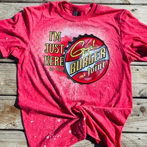 Könnte beinhalten: Rotes T-Shirt mit einem weißen, mit Bleichmittel bespritzten Design. Das Shirt zeigt eine rote, weiße und gelbe Grafik eines Flaschenverschlusses mit dem Text "Guy's Burger Joint Est. 1968" und dem Text "I'm Just Here for the".