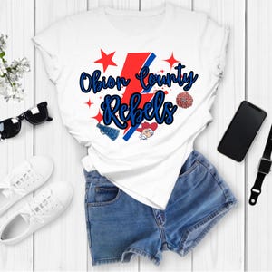 Puede incluir: Camiseta blanca con un diseño gráfico rojo, blanco y azul que dice "Obion County Rebels". El gráfico incluye un rayo, una estrella, un pompón de animadora y un megáfono.