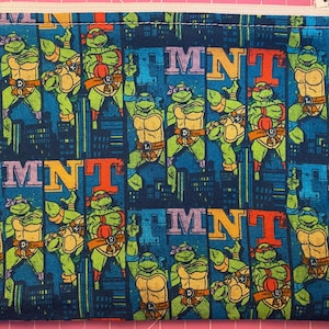 Puede incluir: Una bolsa rectangular con cremallera con un patrón repetido de las Tortugas Ninja en varias poses de acción. El fondo es azul oscuro con siluetas de ciudades y las letras "MNT" en naranja y azul.