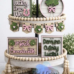 May include: A tiered tray with St. Patrick's Day themed wooden decorations. The decorations include signs with phrases like "How Lucky Are We" and "March Seventeenth", along with shamrocks and bows. The tray is decorated with wooden beads and tassels.