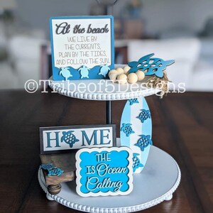 May include: A two-tiered white tiered tray with a blue and white surfboard, a wooden sign that says "HOME" with a sea turtle, a blue sign that says "The Ocean is Calling" with a sea turtle, and a blue sign that says "At the beach we live by the currents, plan by the tides, and follow the sun." with three sea turtles.
