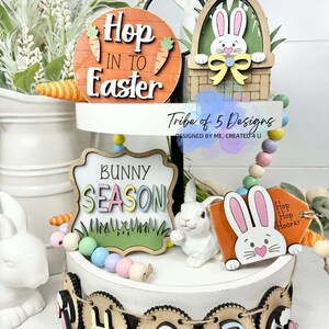 May include: A tiered Easter-themed display with wooden signs and decorations. The signs read "Hop into Easter" and "Bunny Season." A small white bunny figurine and a banner that says "HOP" are also included. The display is decorated with pastel-colored beads.
