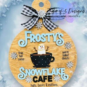 May include: A round wooden sign with the words "Frosty's Snowflake Cafe" in blue and white lettering. The sign features a cartoon snowman holding a cup of hot chocolate with a marshmallow on top. The sign also includes the text "free hot cocoa" and "warm baked donuts".