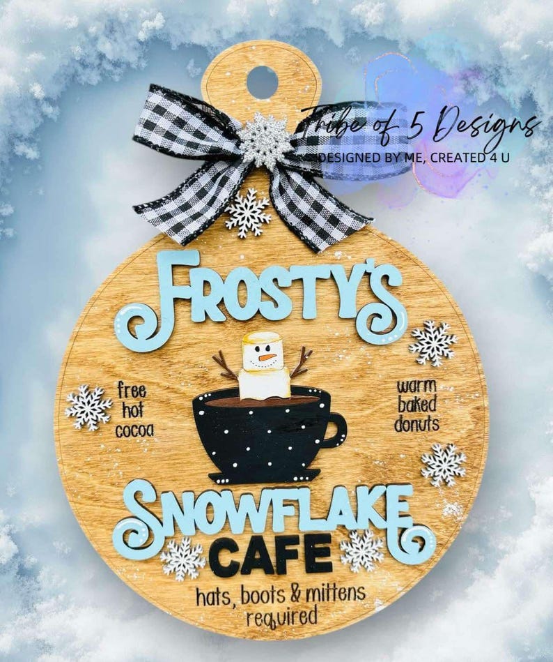 May include: A round wooden sign with the words "Frosty's Snowflake Cafe" in blue and white lettering. The sign features a cartoon snowman holding a cup of hot chocolate with a marshmallow on top. The sign also includes the text "free hot cocoa" and "warm baked donuts".