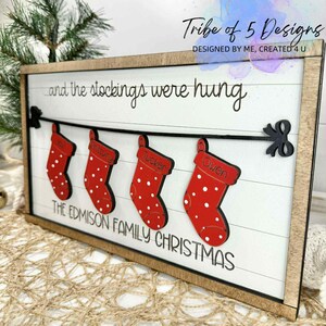 May include: Wooden framed Christmas sign with a white background and the text "...and the stockings were hung." Four red stockings with white polka dots are suspended from a black string. The sign reads "THE EDMISON FAMILY CHRISTMAS."
