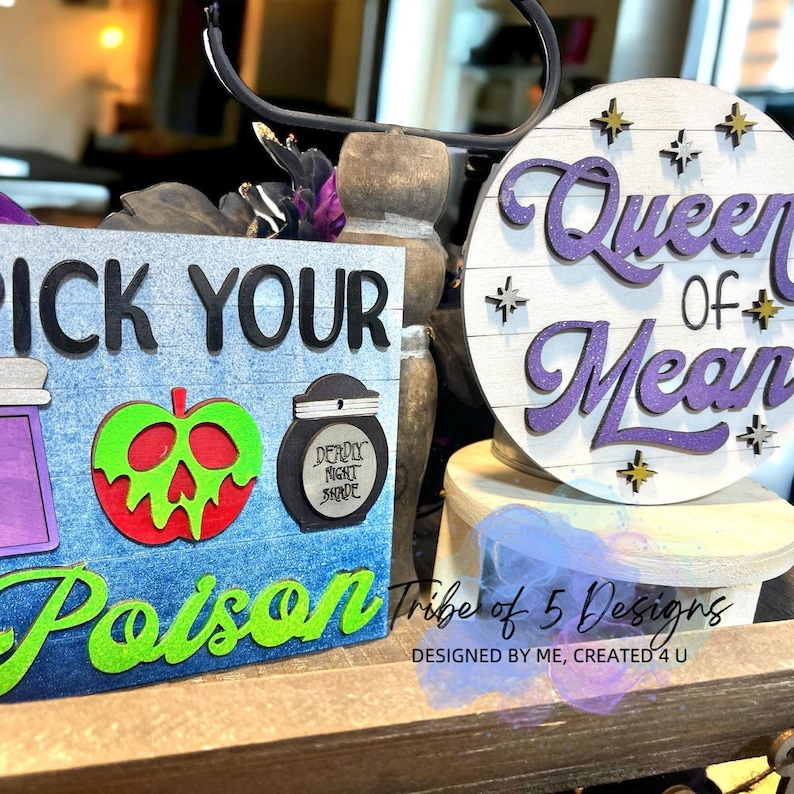 May include: Two wooden signs with a Halloween theme. The first sign is blue with white wood grain and says "Pick Your Poison" with a green apple with a skull face, a purple potion bottle, and a black jar with the text "Deadly Night Shade". The second sign is round and white with purple glitter text that says "Queen of Mean" with gold glitter stars.