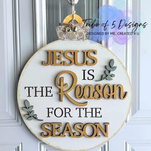 以下が含まれることがあります： 「JESUS IS THE REASON FOR THE SEASON」の文字が茶色と黒で書かれた、丸い白い木製のクリスマスオーナメント。 オーナメントの上部にはベルが付いており、Tribe of 5 Designsによってデザインされています。