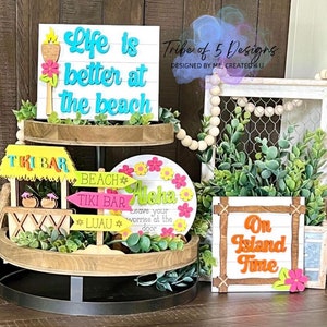 May include: A tiered wooden tray decorated with a beach theme. The top tier features a white sign with the text "Life is better at the beach" in blue and green lettering. The bottom tier features a variety of wooden signs with phrases like "Tiki Bar", "Beach", "Aloha", and "On Island Time".