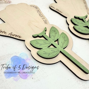 Puede incluir: Dos tarjetas de madera en forma de flor para el D&iacute;a de la Madre con hojas y tallos verdes. Las tarjetas est&aacute;n personalizadas con el texto "Feliz D&iacute;a de la Madre", "Con amor, Oliver", y "Con amor, Aurora".