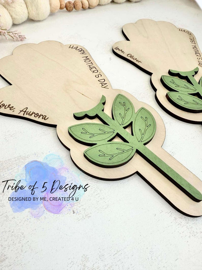Puede incluir: Dos tarjetas de madera en forma de flor para el D&iacute;a de la Madre con hojas y tallos verdes. Las tarjetas est&aacute;n personalizadas con el texto "Feliz D&iacute;a de la Madre", "Con amor, Oliver", y "Con amor, Aurora".