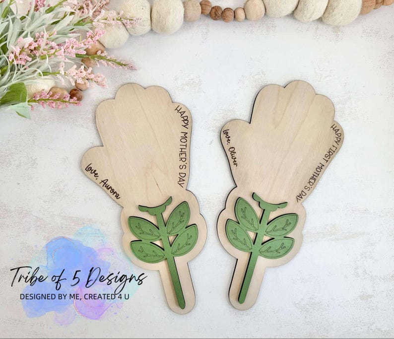 Puede incluir: Dos adornos de madera en forma de mano con dise&ntilde;os florales verdes y el texto "Feliz D&iacute;a de la Madre" y "Feliz Primer D&iacute;a de la Madre". Los adornos est&aacute;n personalizados con los nombres "Aurora" y "Oliver".