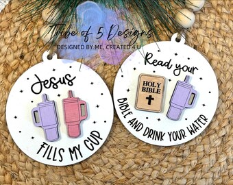 May include: Two round wooden ornaments with white backgrounds and black text. The first ornament says "Jesus fills my cup" with two purple and pink cups with straws. The second ornament says "Read your Bible and drink your water" with a small wooden Bible and a purple cup with a straw.