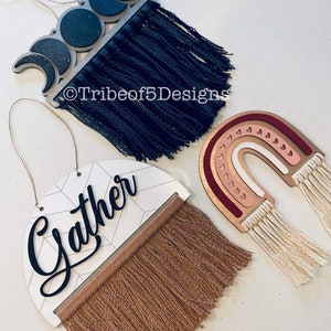 May include: Three wooden wall hangings with fringe. The first is a crescent moon design with black and white paint. The second is a rainbow design with pink, red, and white paint. The third is a half-circle with a geometric pattern and the word "Gather" in black lettering.