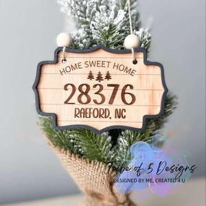 Puede incluir: Un letrero de madera de 'Home Sweet Home' con el texto 'HOME SWEET HOME', el número '28376' y 'RAEFORD, NC'. El letrero está colgado de un pequeño árbol de Navidad artificial.