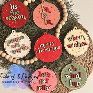 May include: Five wooden Christmas decorations with different festive sayings. The decorations are red, pink, green, and beige with gold glitter accents. The sayings include "Tis the season", "Peace on Earth", "Fa La La", "Merry Christmas", "Warm Wishes", "Let's Get Jolly", and "Oh What Fun".