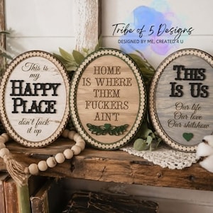 May include: Three oval wooden signs with humorous quotes. The signs have a distressed look with white, green, and gray backgrounds. The text includes phrases like "Happy Place" and "This Is Us". Decorative beads and greenery are also present.