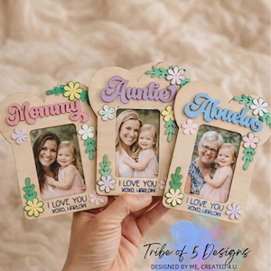 May include: Three wooden heart-shaped photo frames with floral accents. Each frame has a different title: "Mommy", "Auntie", and "Abuela". The frames hold photos of a woman and a young child, with the text "I love you XOXO, Harlow".