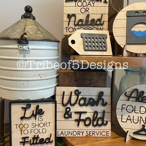 May include: A selection of wooden laundry signs with black lettering. One sign says "Laundry Today or Naked Tomorrow." Another reads "Wash & Fold Laundry Service." A third sign states "Life is too short to fold fitted sheets."