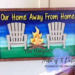 May include: A wooden sign with the text "Our Home Away From Home" and "The Haugen's Est. 2015". The sign features a painted scene of a campfire with two chairs in front of it, against a blue and green background.