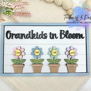 Peut inclure: Panneau en bois avec l'inscription "Grandkids in Bloom" en lettres noires. En dessous, quatre découpes de fleurs avec les noms : Emma, Nalah, Leyla et Lunah. Le panneau a une bordure bleu clair et un fond blanc.
