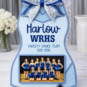 May include: Light blue decorative sign featuring a photo of a dance team. The sign has a bow at the top and text that reads "Harlow WRHS Varsity Dance Team 2022-2026". The bow and text are in a darker shade of blue.
