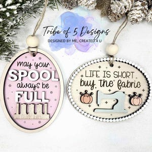Puede incluir: Dos adornos ovalados de madera. Uno dice "may your SPOOL always be FULL" con un diseño de carrete. El otro dice "LIFE IS SHORT buy the fabric" con una máquina de coser y calabazas.
