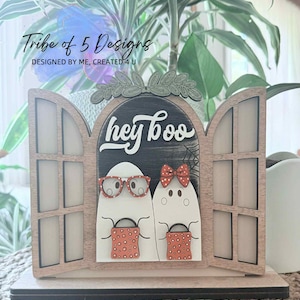 Könnte beinhalten: Halloween-Dekoration aus Holz mit zwei Geisterfiguren, die Süßigkeitentüten halten, eingerahmt von einem Fensterdesign. Der Schriftzug "hey boo" ist über den Geistern zu sehen. Die Dekoration ist aus Holz und hat eine natürliche Oberfläche.