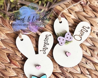 May include: Two wooden bunny-shaped ornaments with the names "Jameson" and "Quinn" written on them. The ornaments are decorated with pink and blue bows, hearts, and a purple flower.