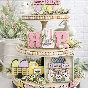 Könnte beinhalten: Ein dreistufiges Osterdekorations-Tablett. Die oberste Ebene zeigt ein Holzschild mit der Aufschrift "Easter Egg Hunt" mit Pastell-Eiern und einem weißen Hasen. Die mittlere Ebene hat das Wort "HOP" und einen Hasen mit Brille. Die untere Ebene hat einen gelben LKW mit Eiern und den Worten "hello PEEPS".