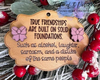 May include: Wooden plaque with the text "TRUE FRIENDSHIPS ARE BUILT ON SOLID FOUNDATIONS." and the phrase "Such as alcohol, laughter, sarcasm, and a dislike of the same people." Features pink bow accents and a hole for hanging.