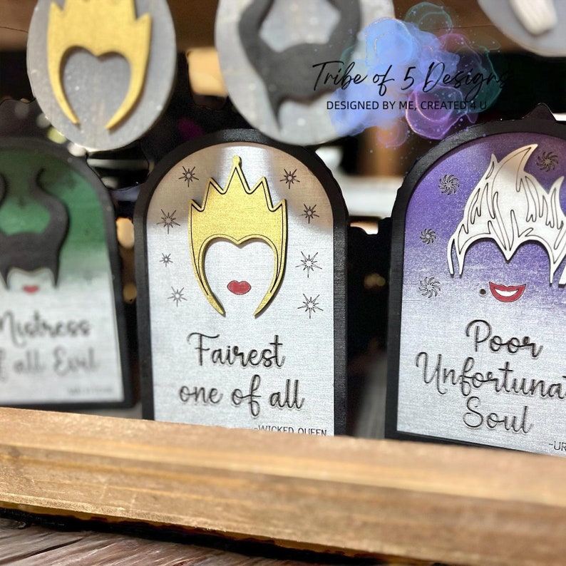 May include: Three decorative wooden signs with silver backgrounds and black borders. Each sign features a different Disney villain: Maleficent, the Evil Queen, and Ursula. The signs have text that reads "Mistress of all Evil", "Fairest one of all", and "Poor Unfortunate Soul".