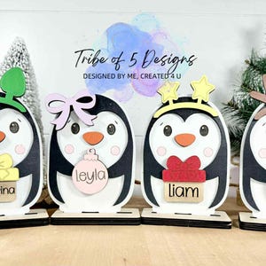 May include: Four wooden penguin decorations with black and white bodies, each with a unique accessory: green ears, a pink bow, yellow stars, and brown antlers. Each penguin has a name tag: Katarina, Leyla, Liam, and Gage.